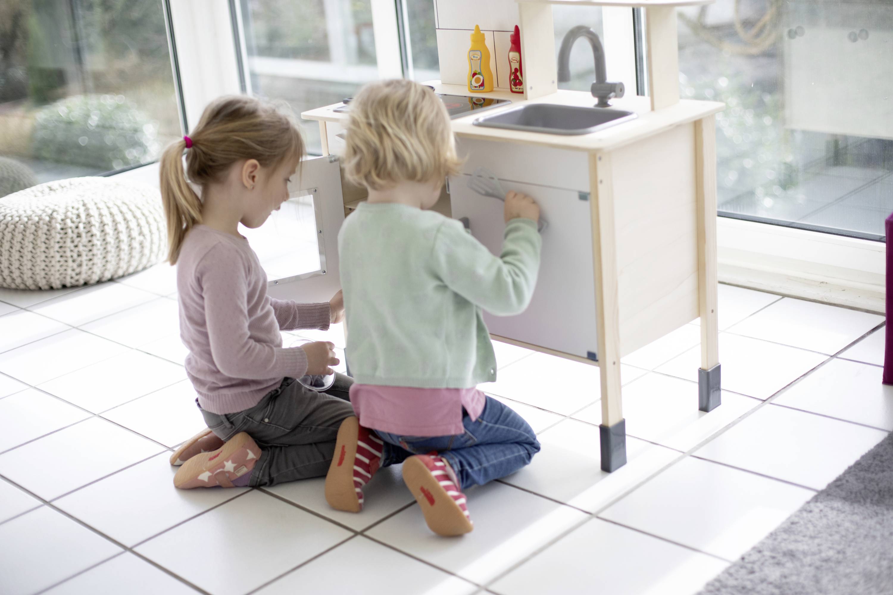 Zwei Kinder spielen mit einer Spielküche. Sie sitzen auf einem weißen Fliesenboden und beschäftigen sich mit den Schranktüren.