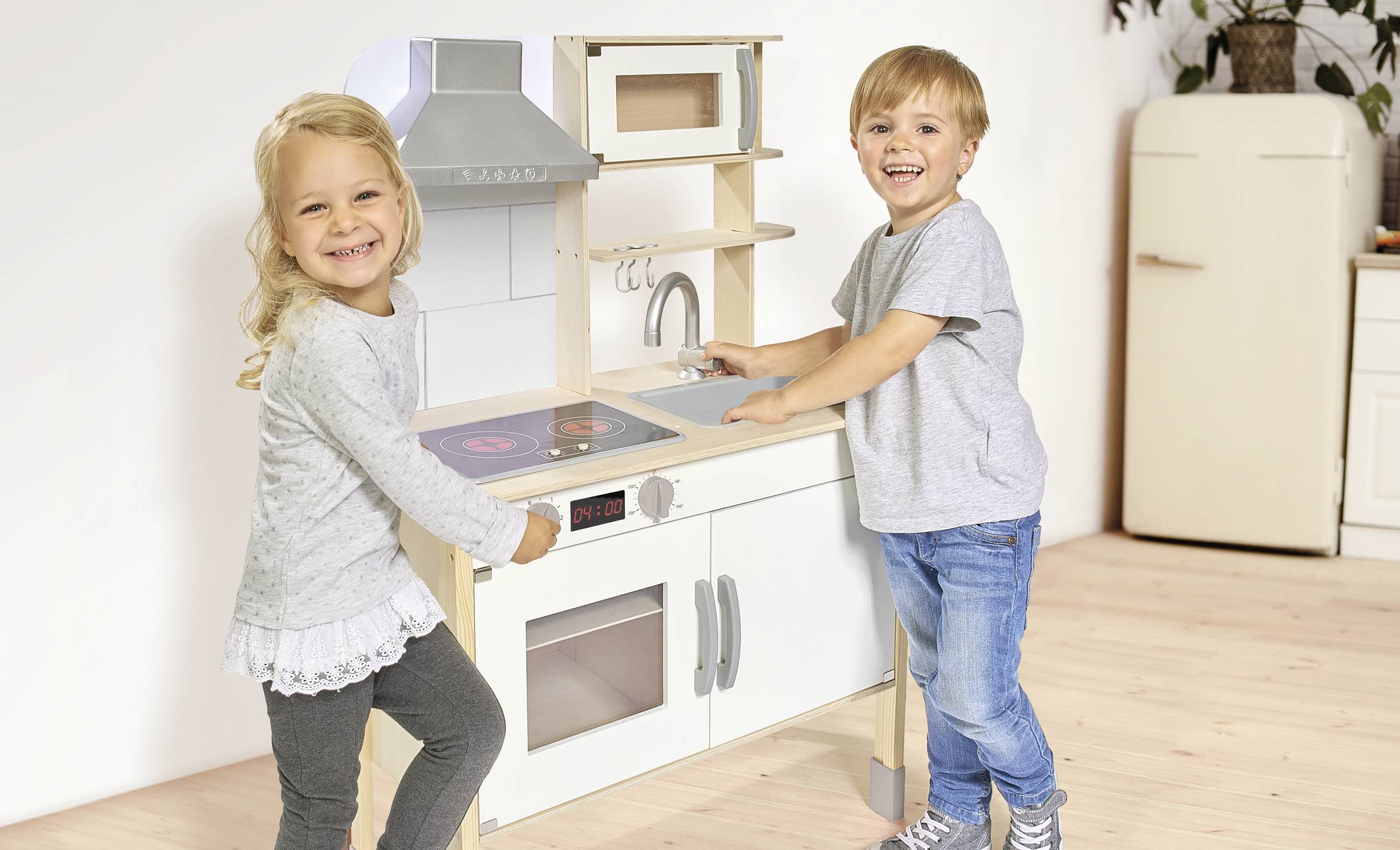Ein Junge und ein Mädchen spielen fröhlich in einer Spielküche aus Holz. Beide lächeln und tragen legere Kleidung.