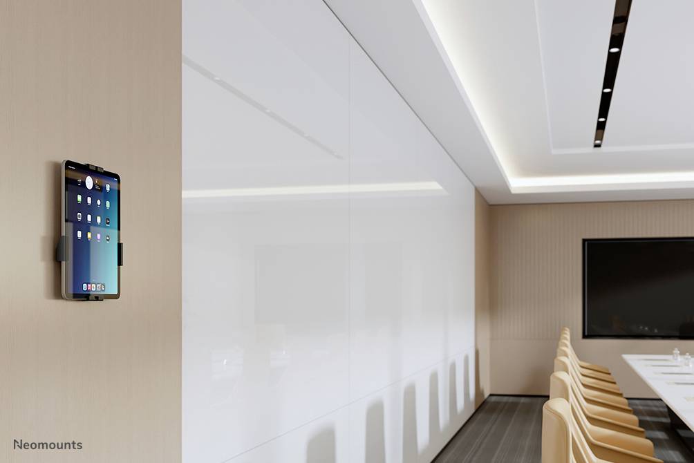 Tablet an einer weißen Wand in einem Konferenzraum mit einem langen Tisch und mehreren Stühlen. An der Wand hängt ein großer Bildschirm.