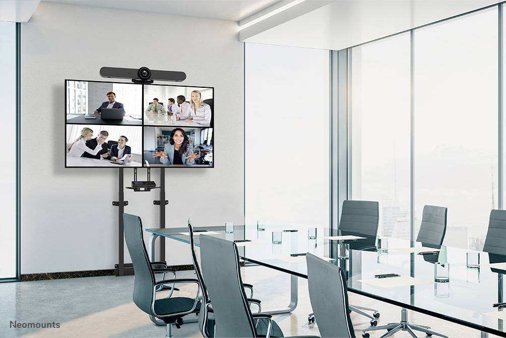 Moderner Konferenzraum mit Glastisch und Stühlen, ein großer Bildschirm zeigt ein Videomeeting mit fünf Personen in unterschiedlichen Ansichten.