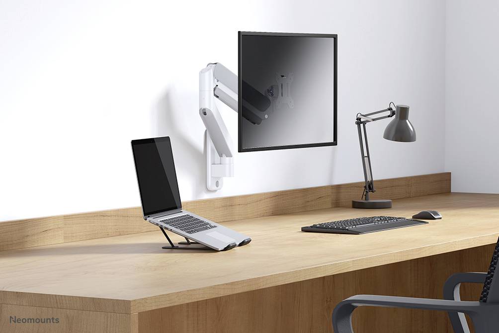 Ein Schreibtisch mit einem Laptop auf einem Ständer neben einem Monitor an der Wandhalterung. Eine Lampe und eine Tastatur sind ebenfalls vorhanden.