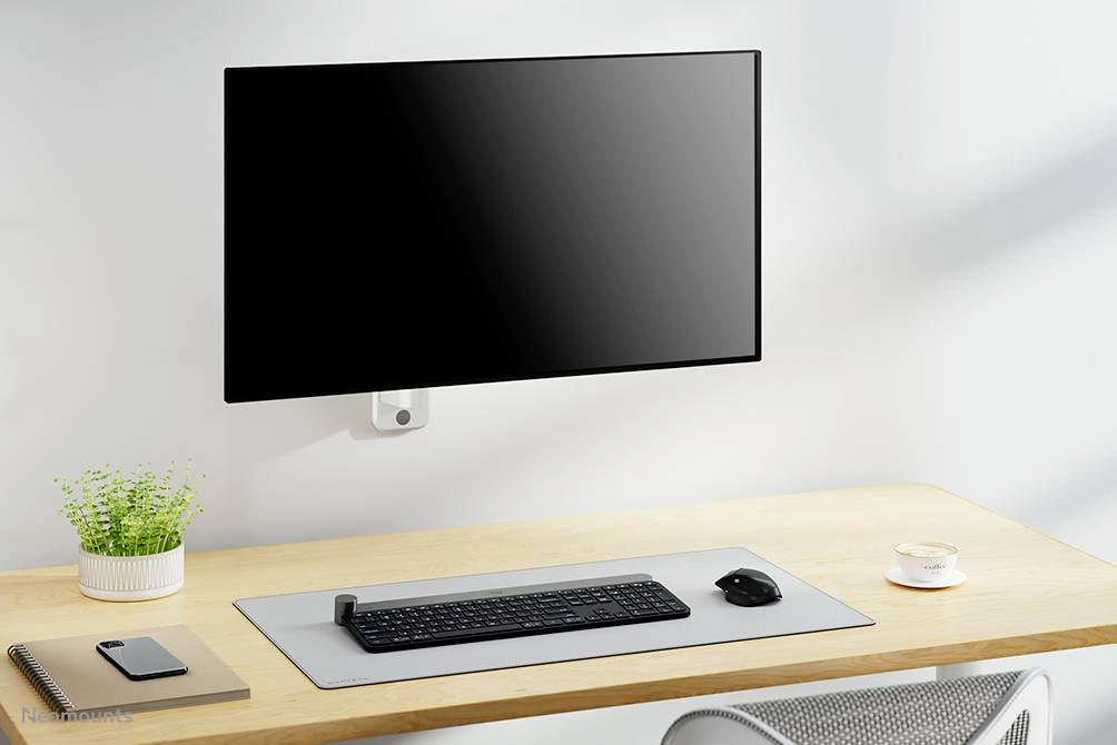 Ein ordentlicher Schreibtisch mit einem Wandmonitor, einer Tastatur und Maus auf einem grauen Mauspad, Notizbuch, Stift und Topfpflanze.