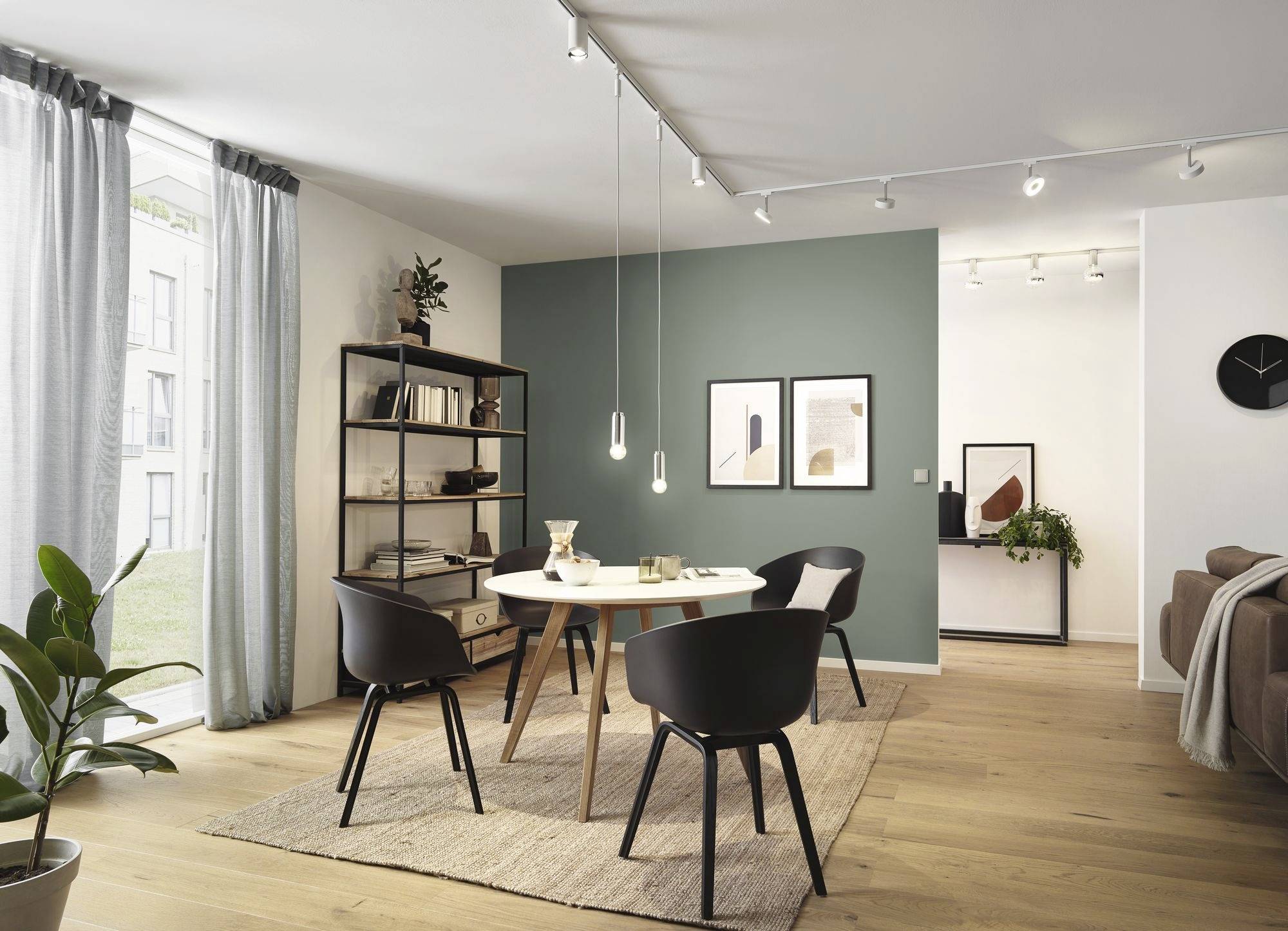 Esszimmer mit rundem Tisch und vier schwarzen Stühlen, Holzboden, graue Akzentwand, Bücherregal, und großen Fenstern mit Vorhängen.