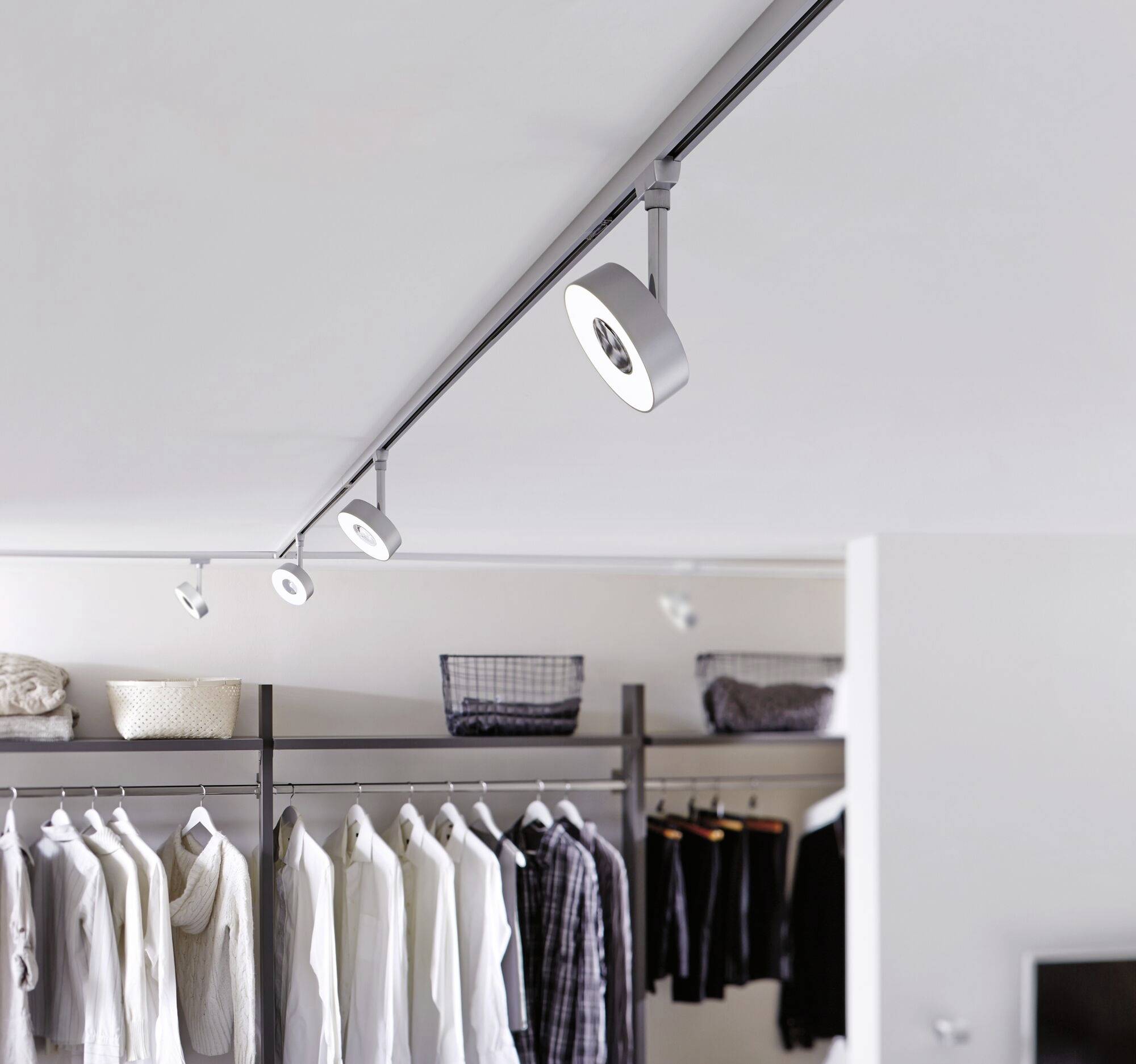 Kleiderschrank mit hängender Kleidung in einem modernen, minimalistisch gestalteten Raum mit Schienensystembeleuchtung an der Decke.