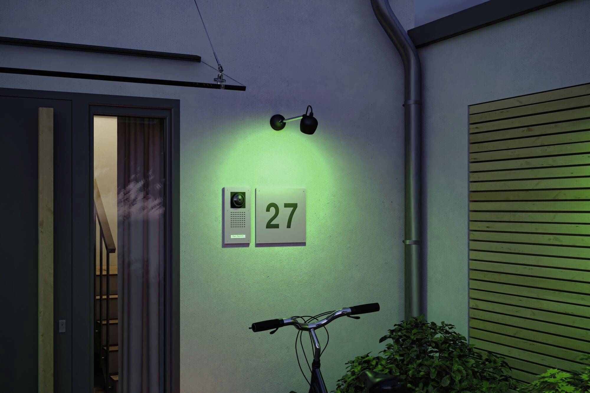 Hauswand bei Nacht mit grüner Beleuchtung, Hausnummer '27' neben einer Gegensprechanlage. Im Vordergrund ist ein Fahrrad vor einem Fenster.