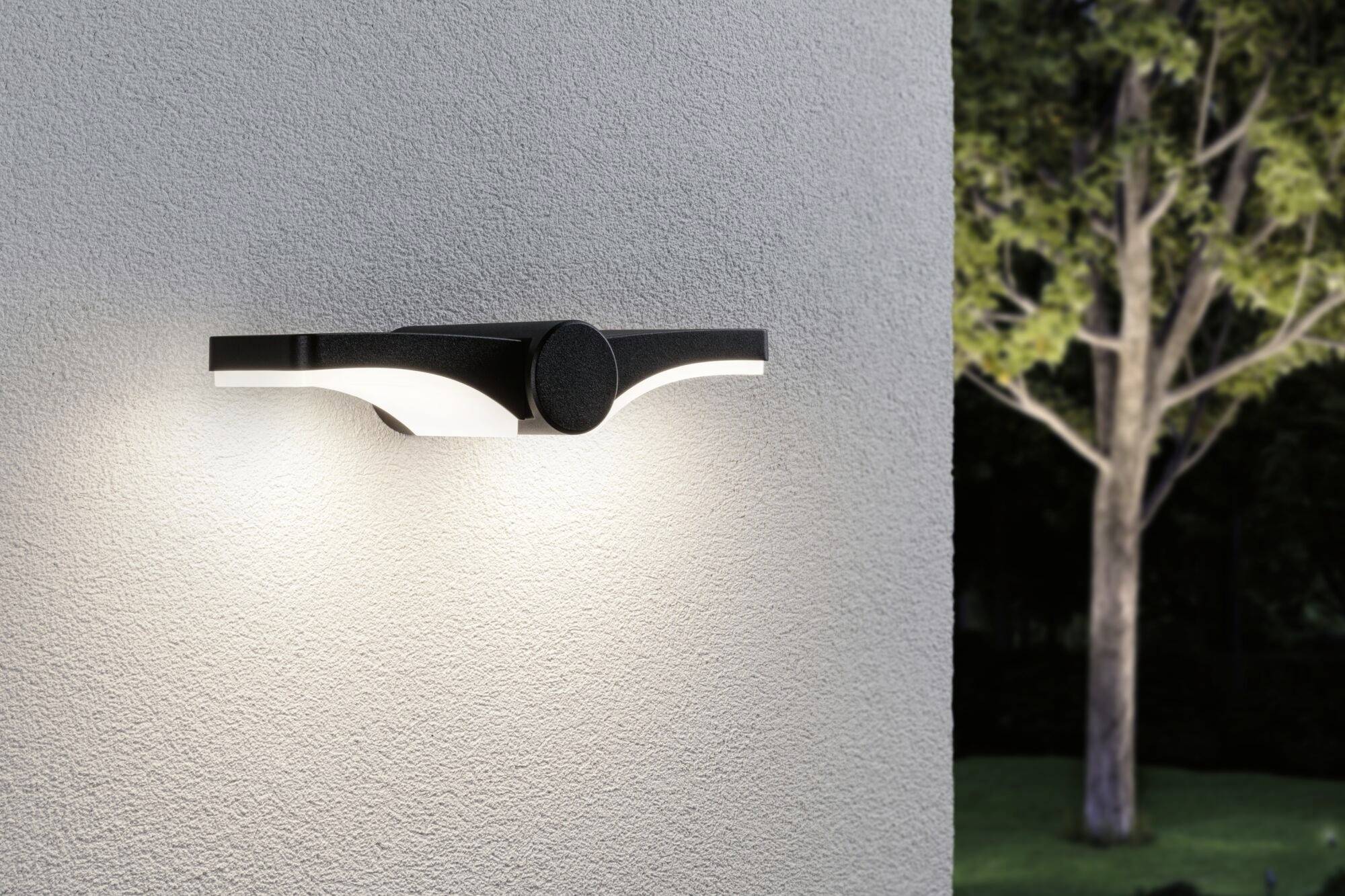 Moderne Wandaußenleuchte an einer hellen Wand montiert, beleuchtet den Bereich darunter. Im Hintergrund ist ein Baum sichtbar.