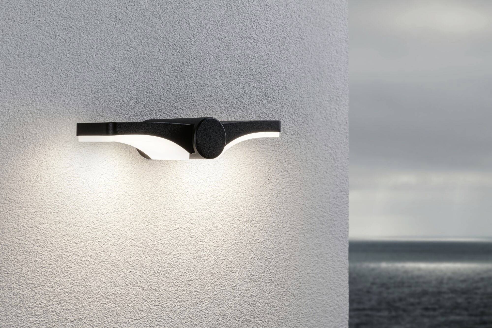 Moderne Wandleuchte an einer weißen Wand, beleuchtet den Außenbereich. Im Hintergrund sieht man das Meer unter einem bewölkten Himmel.