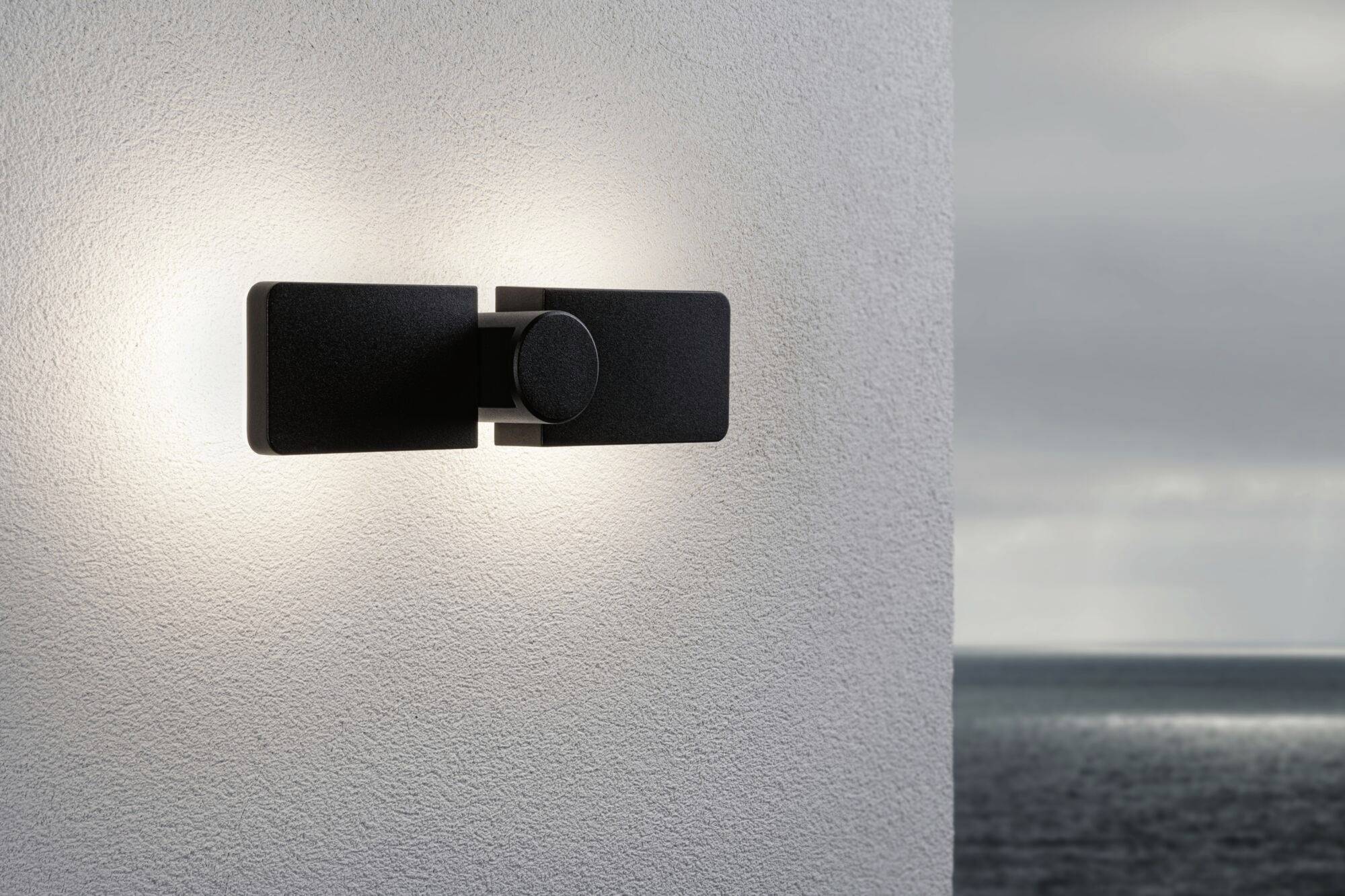 Moderne Außenwandleuchte an einer weißen Wand mit Blick auf ein trübes Meer im Hintergrund. Die Lampe strahlt diffuses Licht aus.