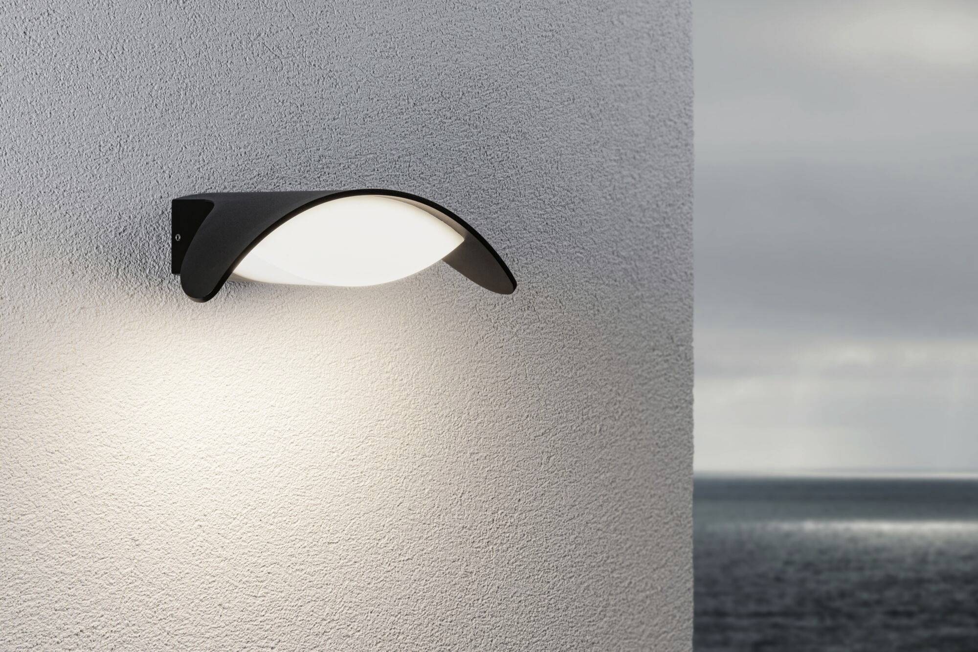 Eine moderne Außenlampe an einer hellen Wand strahlt weiches Licht aus. Im Hintergrund ist das Meer unter bewölktem Himmel zu sehen.