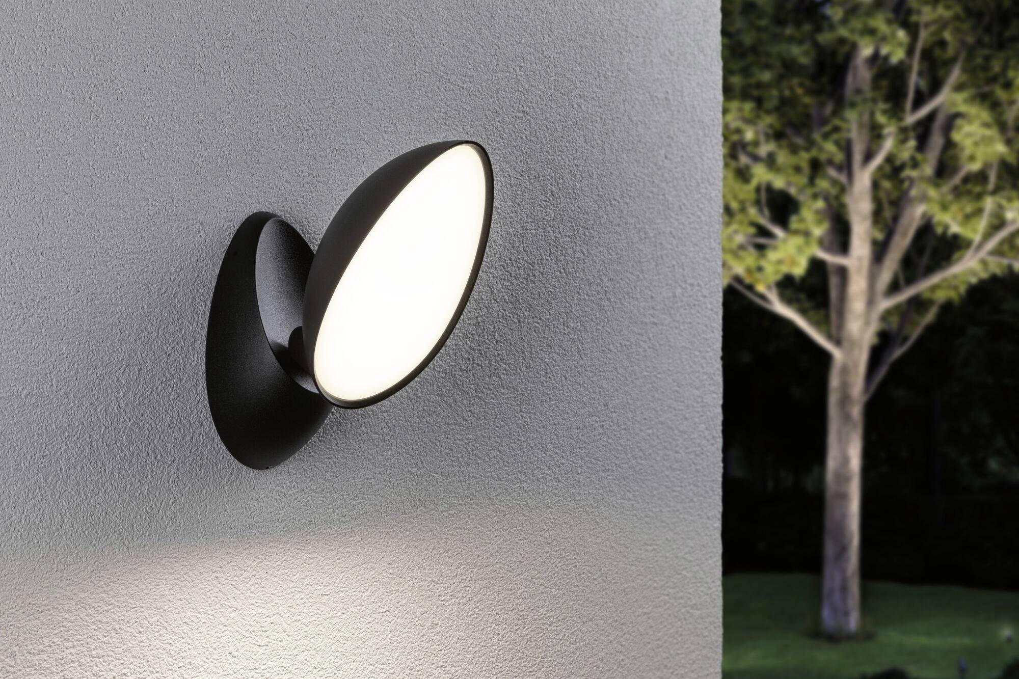 Schwarze Wandleuchte strahlt indirektes Licht gegen eine graue Wand. Im Hintergrund ist ein Baum bei Dämmerung zu sehen.