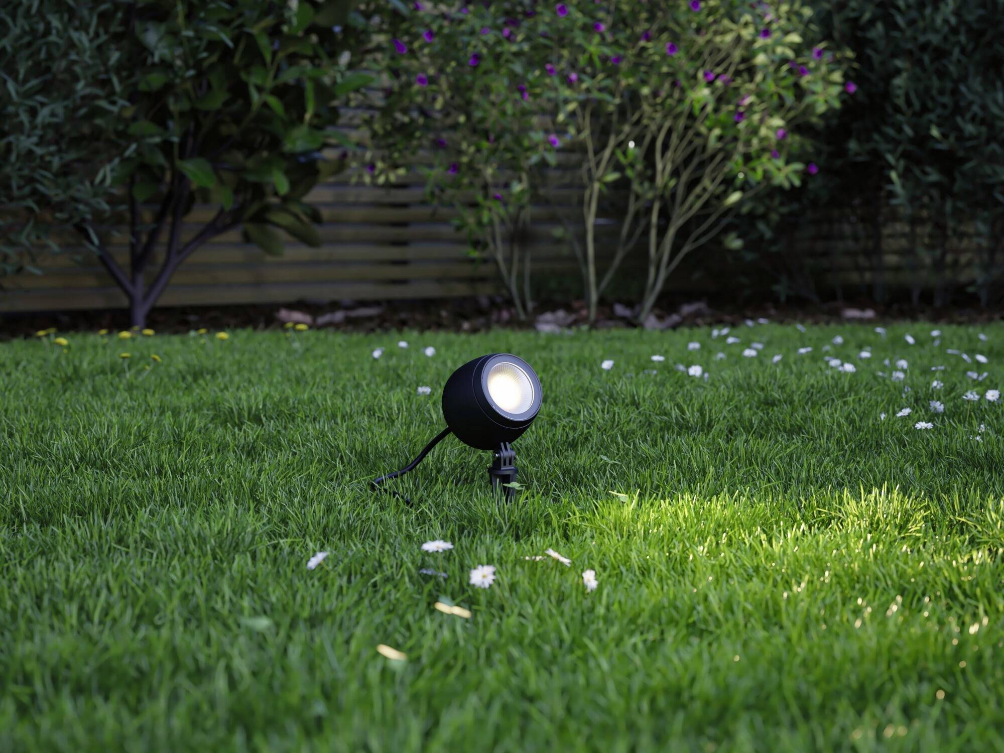Eine schwarze Außenlampe leuchtet auf einem grünen Rasen, umgeben von Blumen und Büschen im Hintergrund.