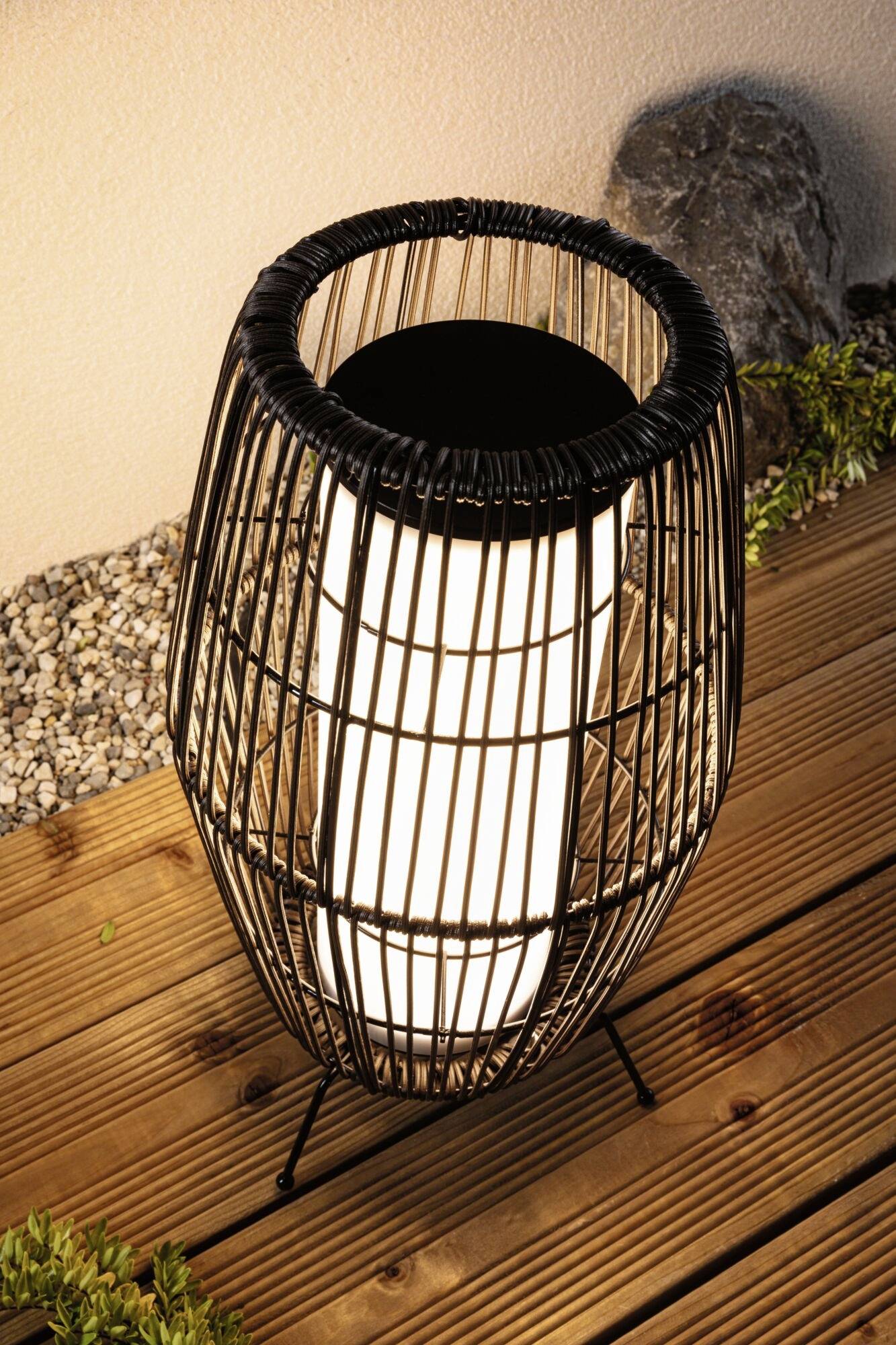 Eine stilvolle Lampe im Freien auf einem Holzboden, umgeben von Kies und Pflanzen, leuchtet sanft in der Dunkelheit.