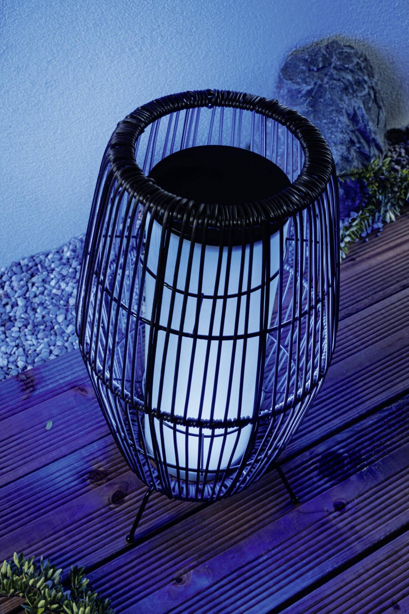 Eine moderne Outdoor-Laterne mit schwarzem Metallkäfig und blauer Hintergrundbeleuchtung auf Holzterrasse, umgeben von Kieselsteinen.