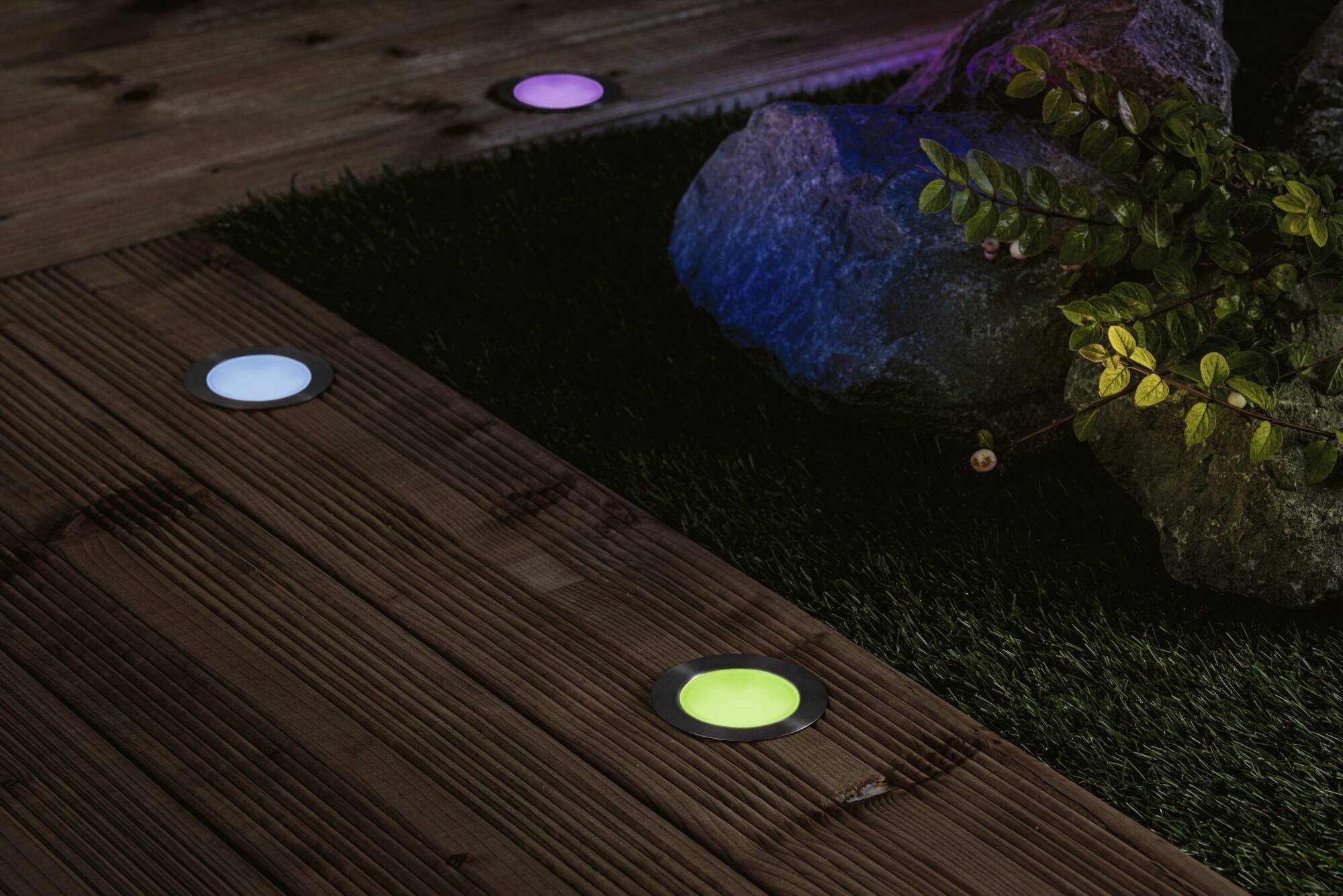 Bunte Bodenlichter auf einer Holzterrasse neben Steinen und Pflanzen, die eine stimmungsvolle Außenbeleuchtung schaffen.