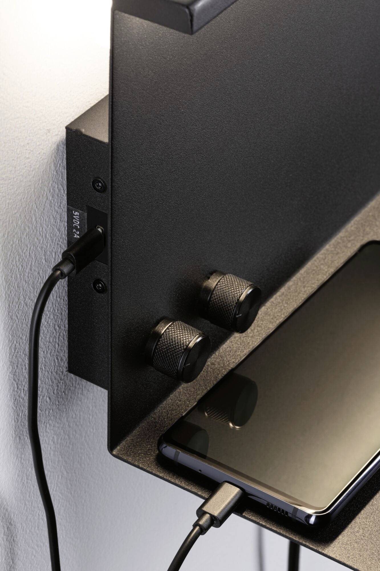 Ein Smartphone, das auf einer schwebenden, schwarzen Ablage liegt, wird über ein Kabel aufgeladen. Daneben sind zwei Drehknöpfe sichtbar.