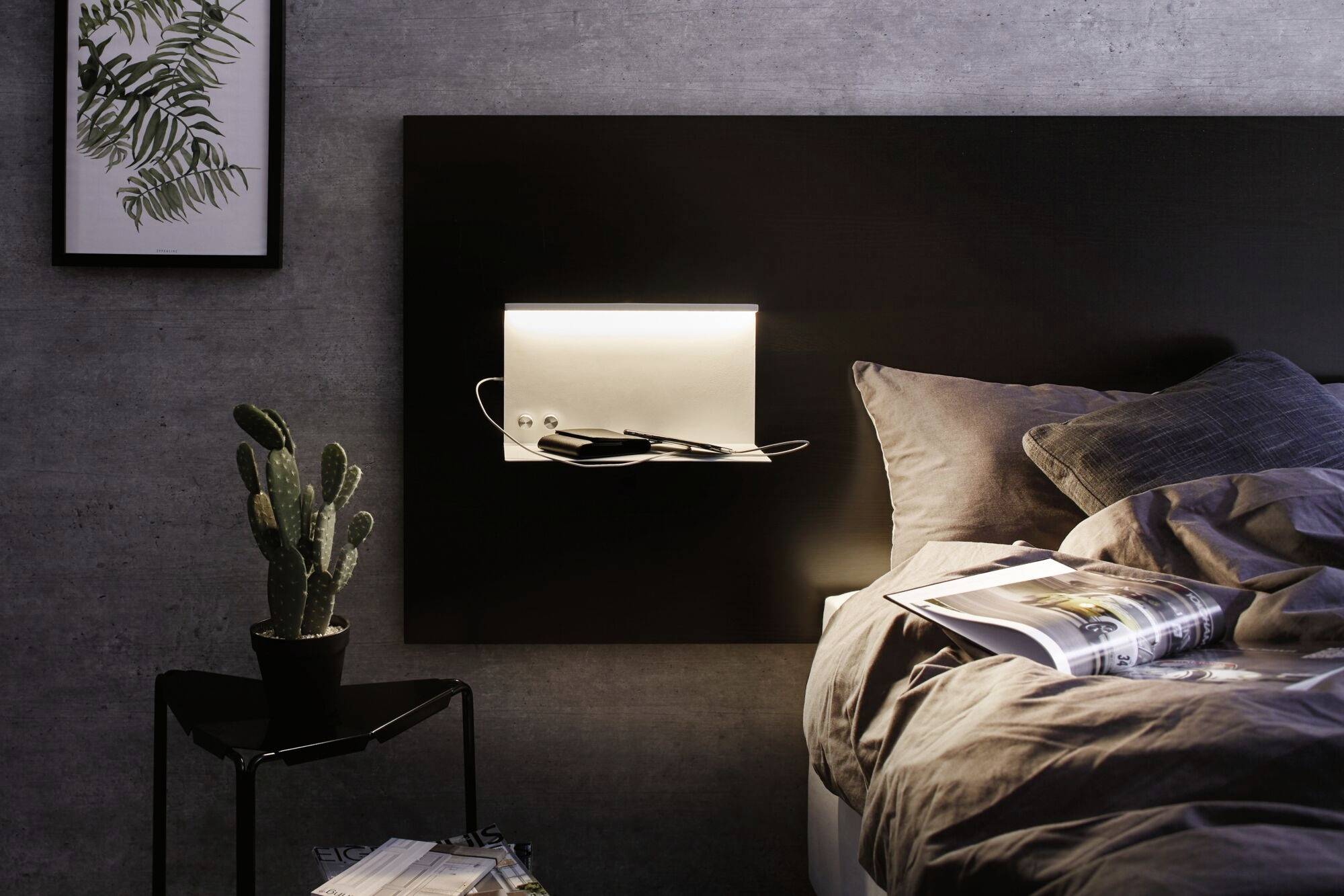 Ein gemütlich eingerichtetes Schlafzimmer mit Bett, darüber ein Regalbrett mit Beleuchtung. Ein Kaktus steht auf einem kleinen Tisch.
