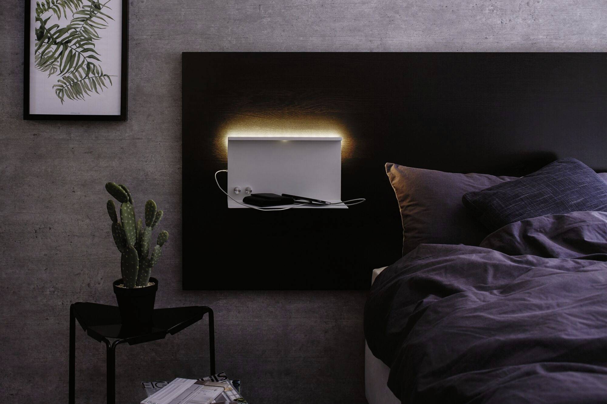 Ein beleuchtetes Wandregal neben einem Bett in einem modernen Schlafzimmer, dekoriert mit einem Kaktus und gerahmtem Pflanzenbild.