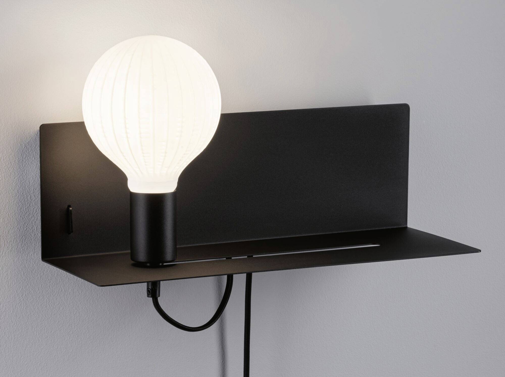 Schwarze Wandleuchte mit integriertem Regal. Runde weiße Glühbirne leuchtet, Kabel hängt herunter. Funktionales modernes Design.