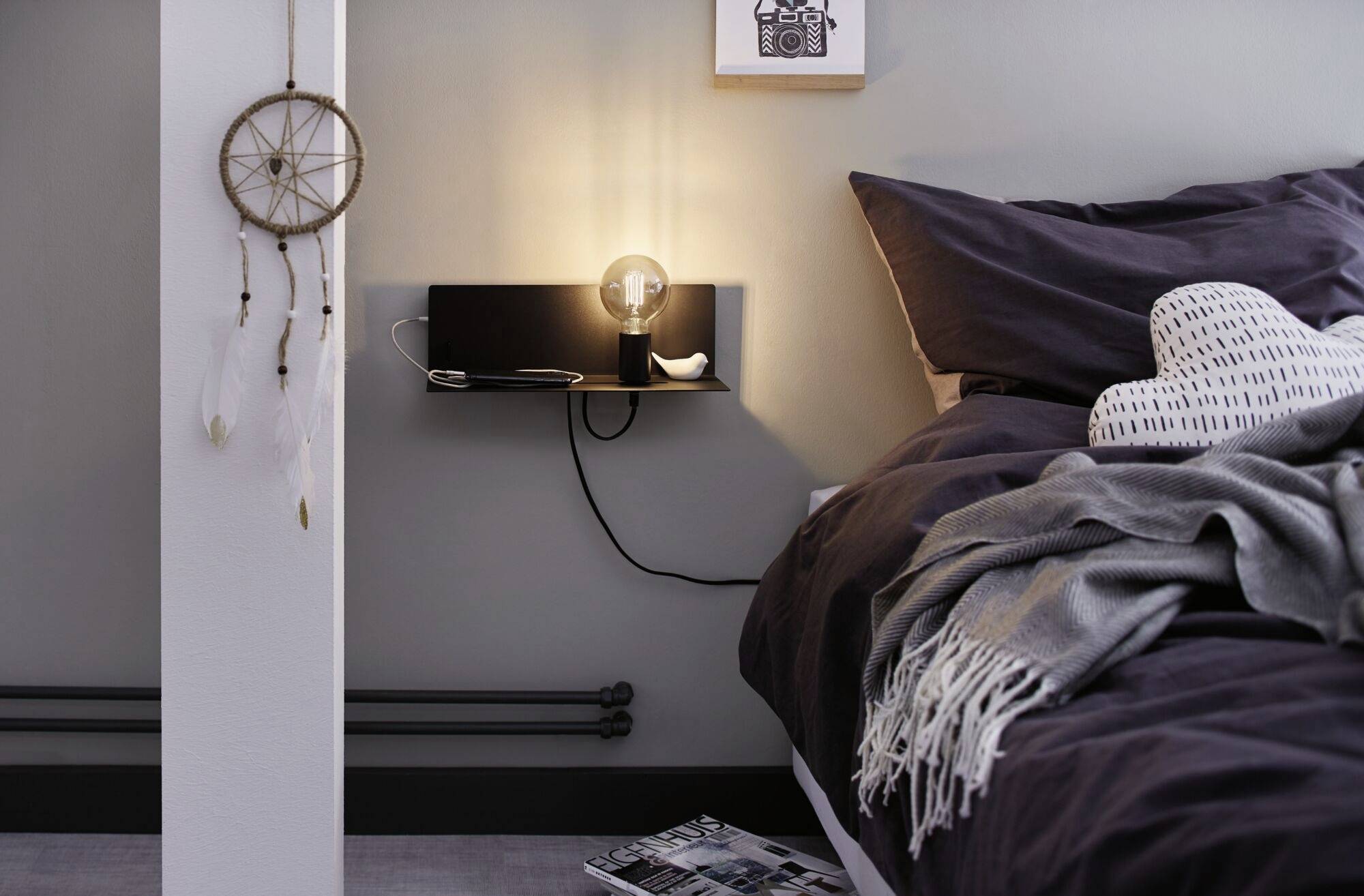 Gemütliches Schlafzimmer: Ein Leuchtmittel auf einem Regal neben einem Bett mit grauer Bettwäsche, Traumfänger an der Wand.