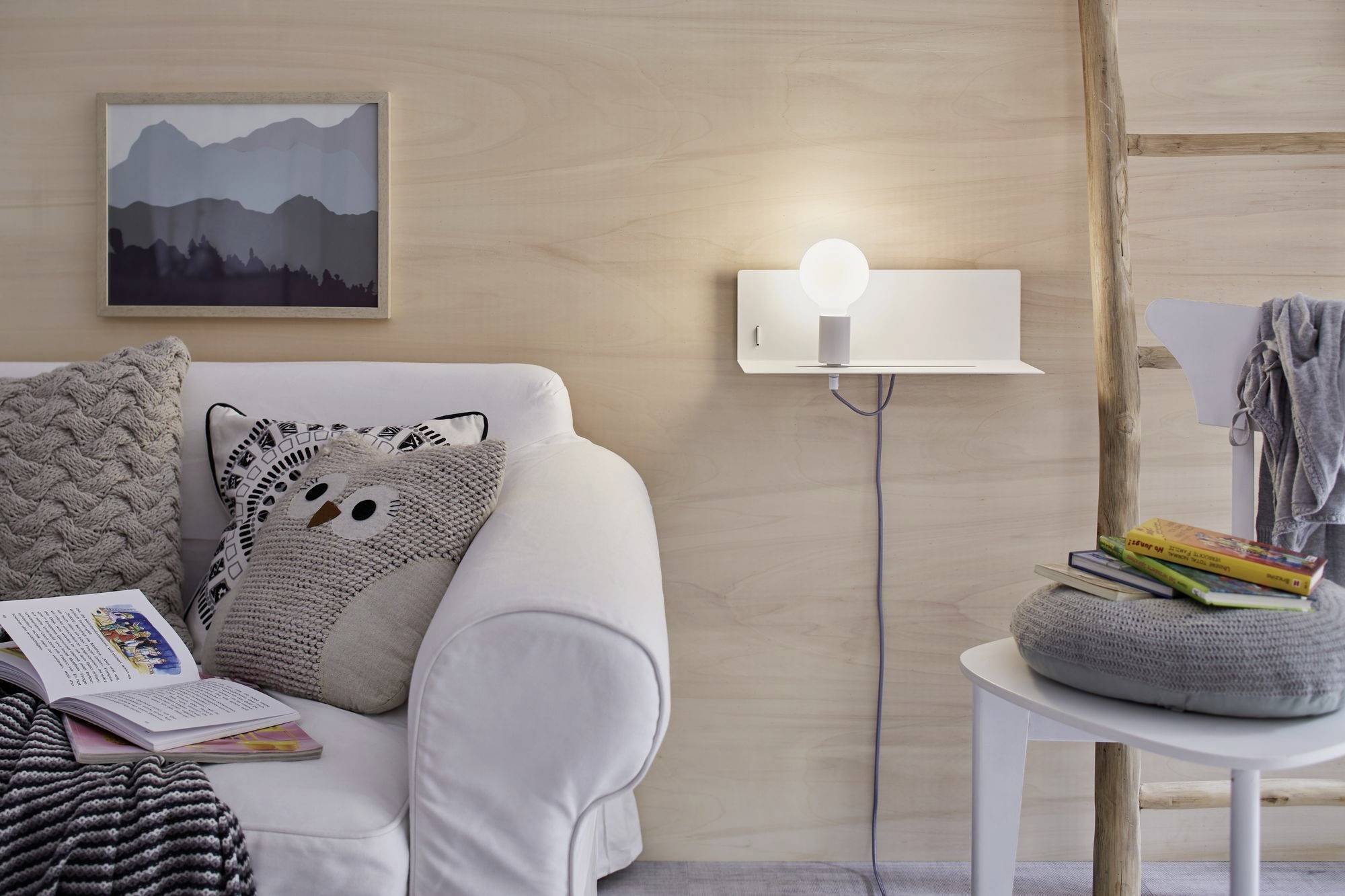 Wohnzimmer mit weißem Sofa, dekorativen Kissen und Decke. Ein Stapel Bücher auf einem Hocker, Wandregal mit Lampe, Holzwand.