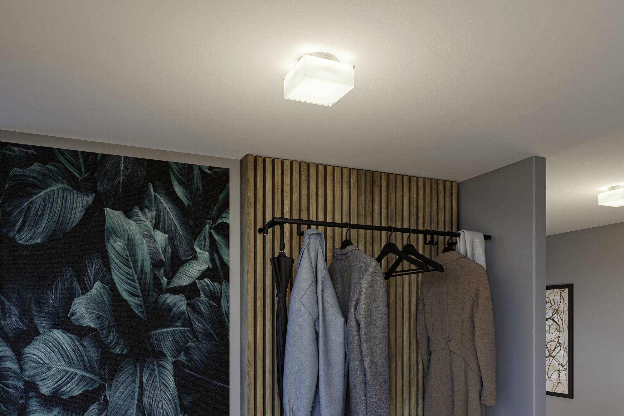 Die Decke zeigt eine moderne Leuchte. Darunter hängt an einer Kleiderstange Kleidung. An der Wand befindet sich eine Tapete mit Blättermuster.