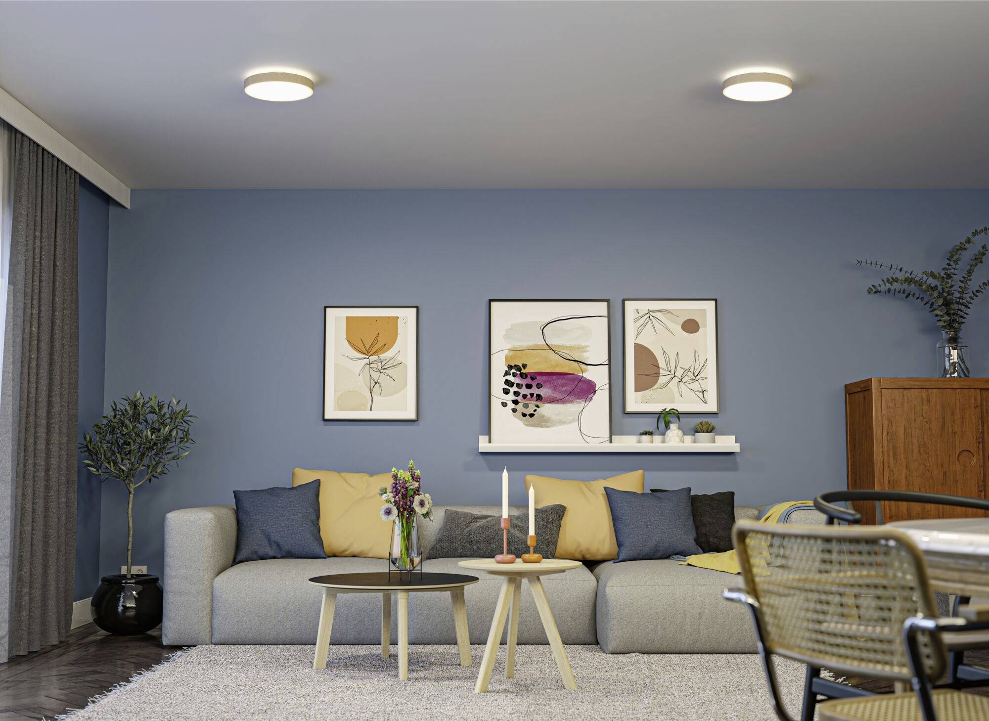 Wohnzimmer mit blauer Wand, grauem Sofa, gelben und blauen Kissen, drei Wandbildern, rundem Couchtisch mit Kerzen und Blumen, Pflanzen, Holzmöbeln.