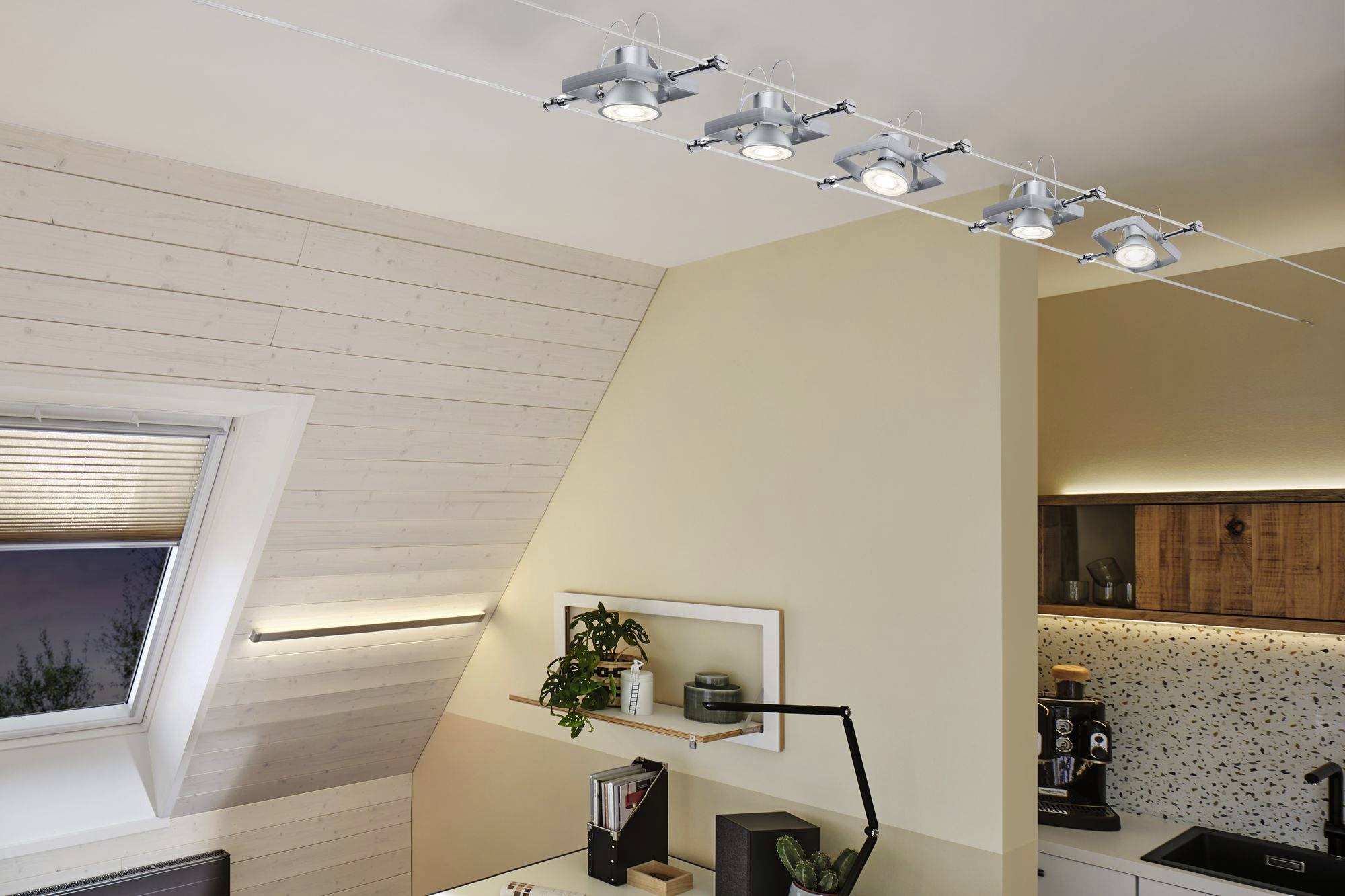 Ein modernes Zimmer mit Schreibtisch, Pflanzen und Regal. An der Decke eine Reihe stilvoller Deckenlampen. Helle und warme Atmosphäre.