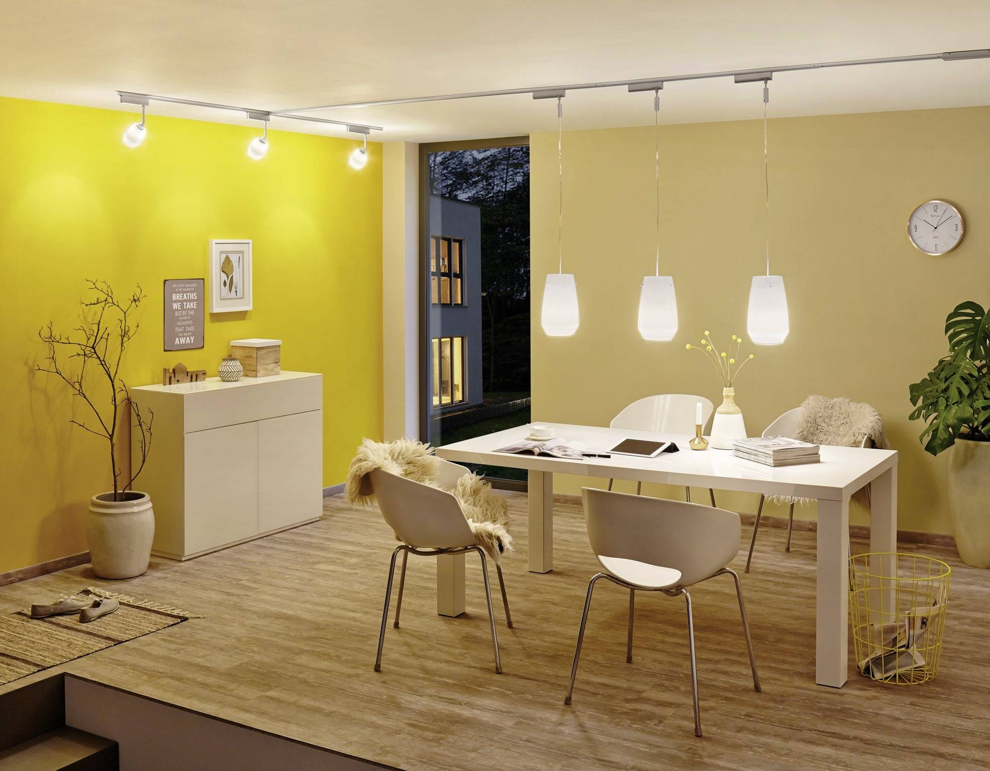 Ein modern eingerichtetes Esszimmer mit gelben Wänden, einem weißen Tisch, Stühlen und hängenden Lampen. Fenster zeigt Abenddämmerung.