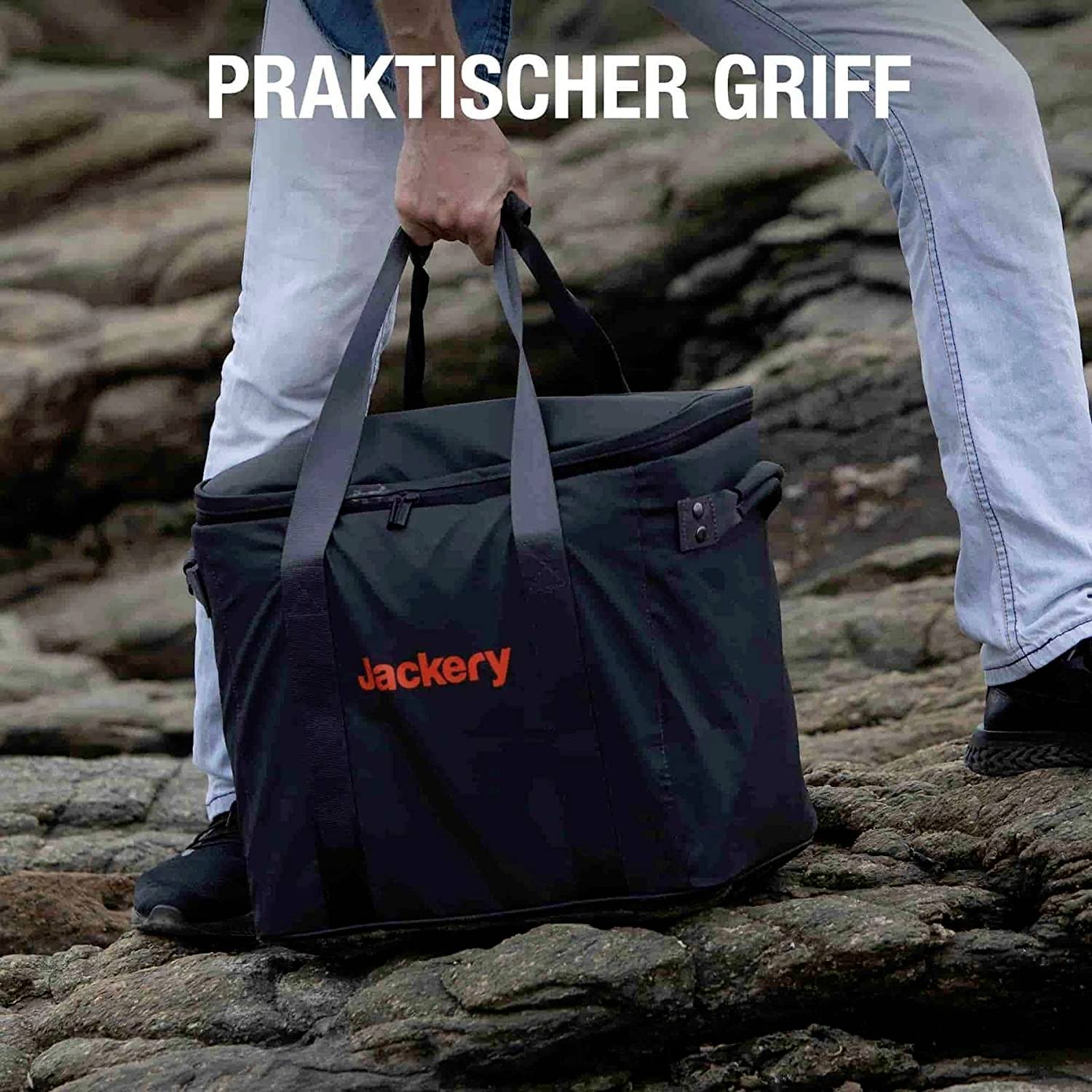 Eine Person trägt eine schwarze Tasche mit 'Jackery'-Logo über unebenem Gelände; der Text 'PRAKTISCHER GRIFF' ist oben zu sehen.