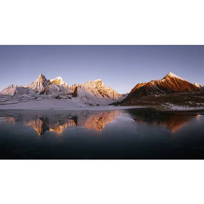 Bergpanorama mit schneebedeckten Gipfeln und ruhigem See im Vordergrund, reflektiert das warme Licht der untergehenden Sonne.