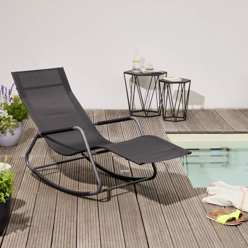 Ein schwarzer Schaukelstuhl steht neben einem Pool auf einer Holzterrasse. Neben dem Stuhl sind zwei kleine Tische mit Getränken.