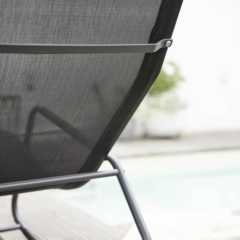 Ein moderner Liegestuhl am Rand eines Pools, im Hintergrund verschwommene grüne Pflanzen.