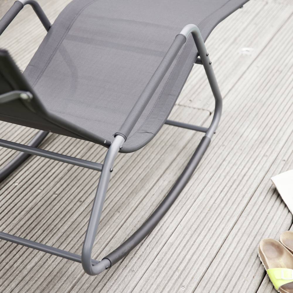 Ein grauer Liegestuhl steht auf einem Holzdeck, Sonnenlicht fällt darauf. Eine Handtasche und Sandalen sind im Bildausschnitt zu sehen.