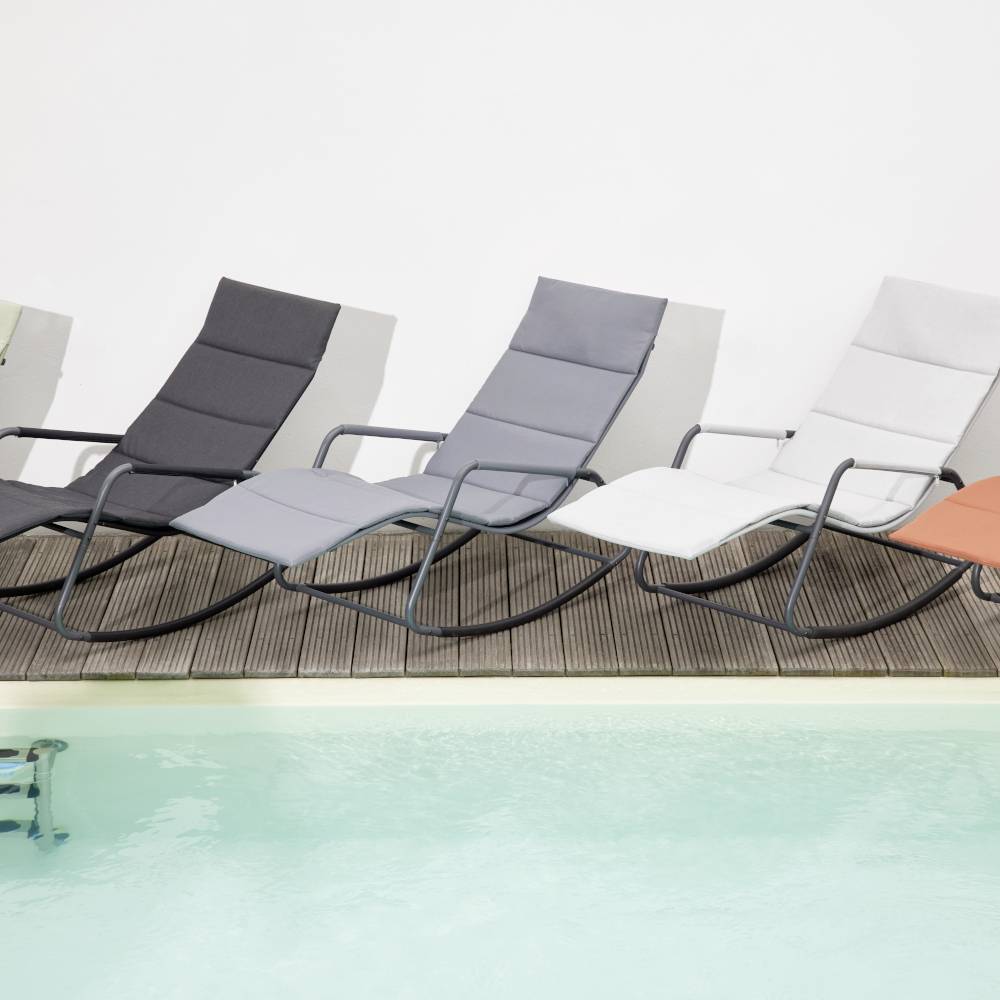 Vier moderne Liegestühle in verschiedenen Grautönen stehen auf einer Holzterrasse neben einem Pool mit türkisfarbenem Wasser.