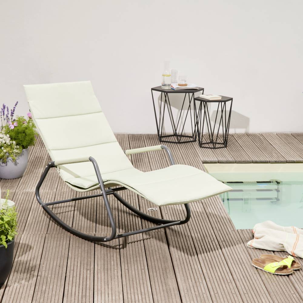 Ein Schaukelstuhl steht auf einer Holzterrasse neben einem kleinen Tisch mit Getränken und Blumenvasen. Im Hintergrund ist ein Poolbereich sichtbar.