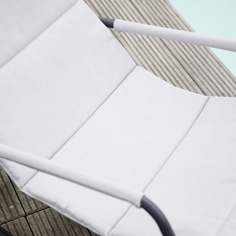 Ein hellgrauer Liegestuhl steht auf einem hölzernen Terrassendeck neben einem Pool mit türkisfarbenem Wasser.