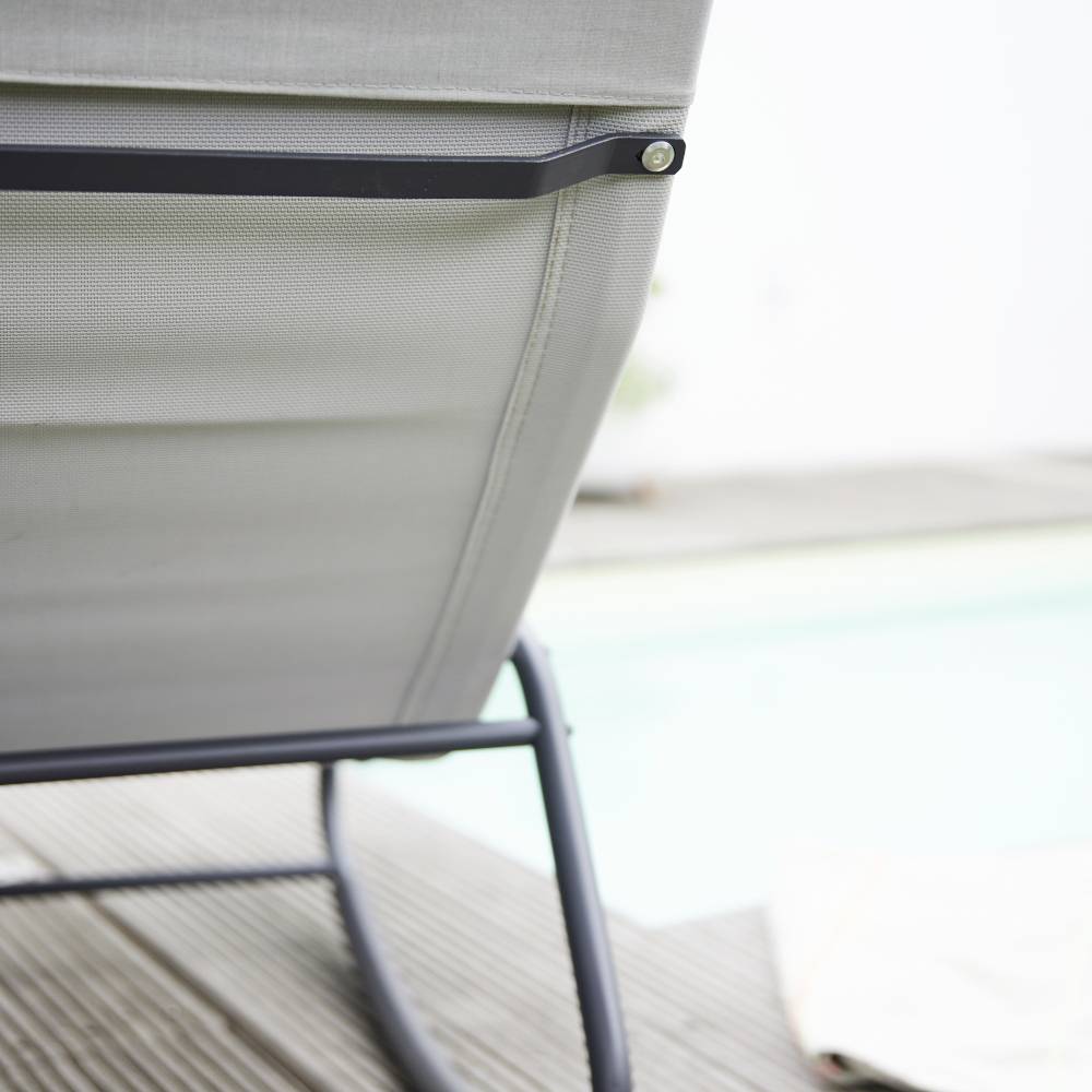 Graumetallene Rückenlehne eines Stuhls auf einer Holzterrasse, links daneben ein verschwommener Pool im Hintergrund.