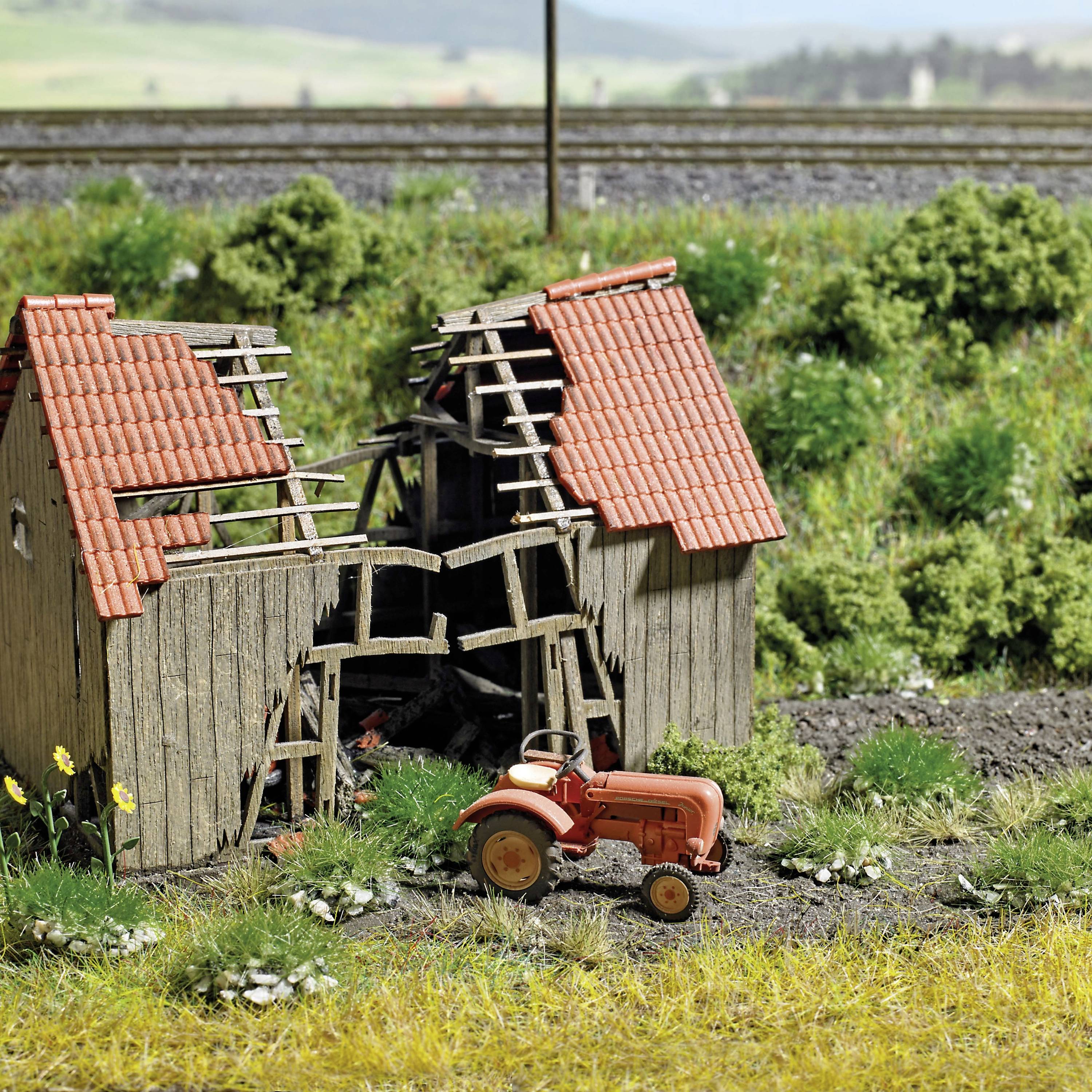 Holzhütte mit eingestürztem Dach und rotem Traktor im Vordergrund; ländliche Szenerie mit Gras und Bahngleisen im Hintergrund.