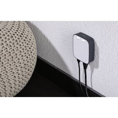 Ein smartes Gerät ist an eine Wandsteckdose angeschlossen. Zwei schwarze Kabel führen von ihm weg, es befindet sich in einem modernen Wohnraum.