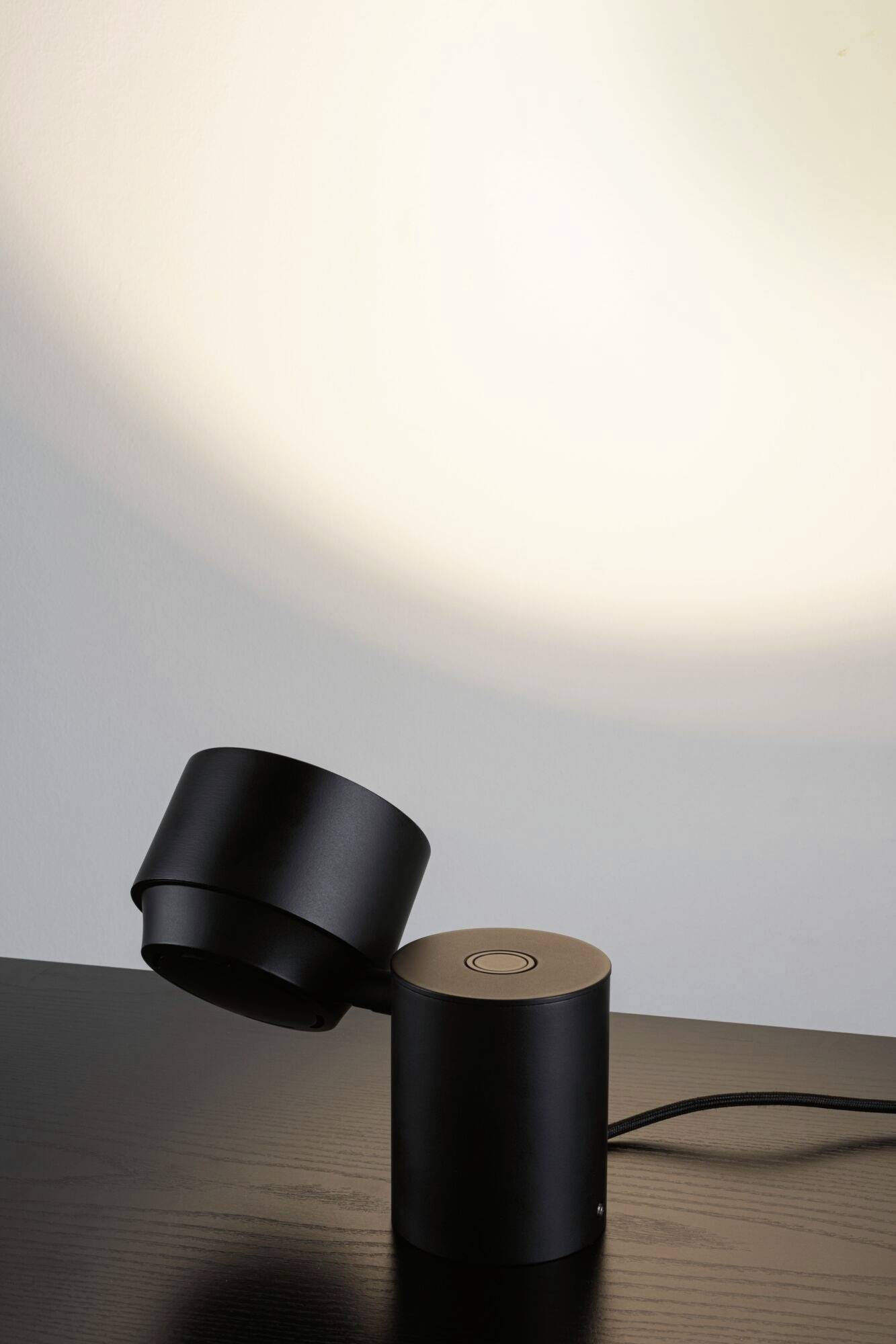 Schwarze Tischleuchte mit modernem Design auf dunklem Holztisch, beleuchtet Wand in sanftem Licht.