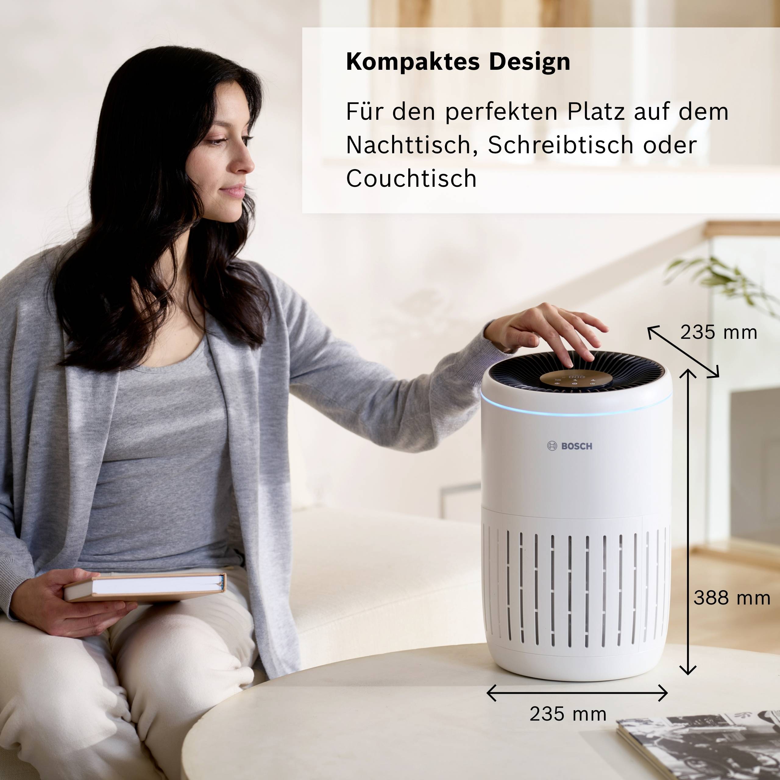 Frau sitzt auf Sofa, hält Buch, daneben Luftreiniger. Text oben rechts: 'Kompaktes Design, ideal für Nacht-, Schreib- oder Couchtisch'.