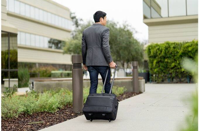 Mann in Business-Kleidung zieht einen schwarzen Rollkoffer auf einem Gehweg zwischen Bürogebäuden entlang.