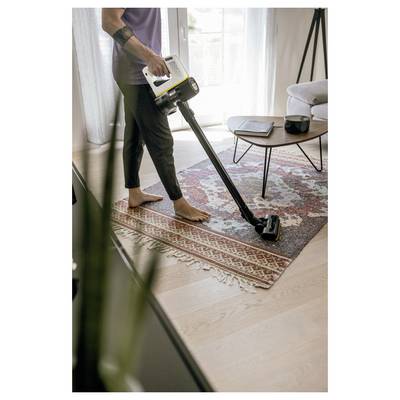 Eine Person saugt einen gemusterten Teppich in einem Wohnzimmer mit einem kabellosen Staubsauger. In der Nähe steht ein kleiner Tisch mit Gegenständen darauf.