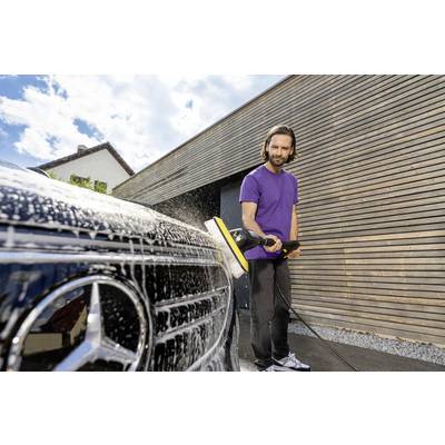 Eine Person in einem lila Hemd reinigt ein schwarzes Auto mit einem Hochdruckreiniger vor einer modernen Holzgarage unter einem teilweise bewölkten Himmel.