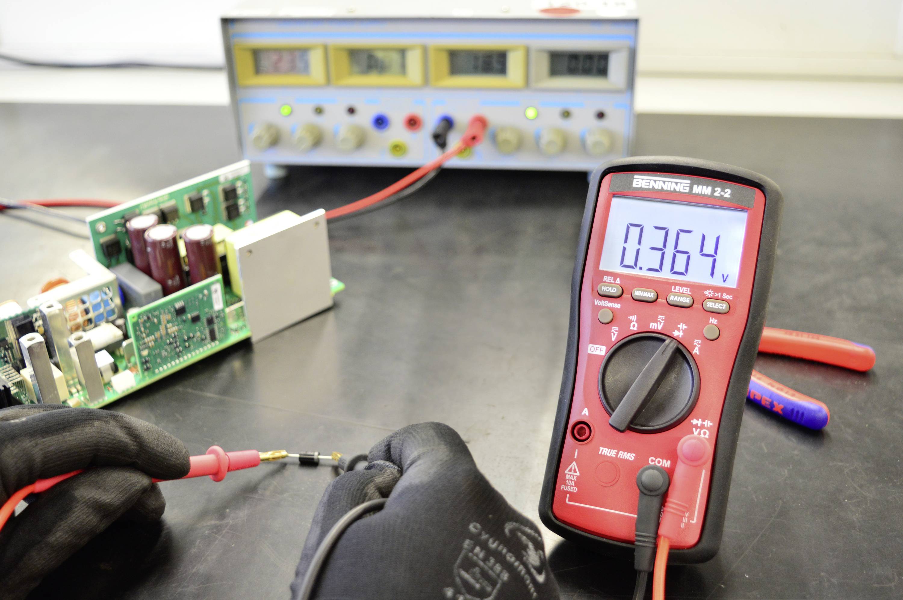 'Ein Multimeter zeigt 0,364 V an, daneben ein geöffnetes Gerät. Im Hintergrund ein Netzgerät und verschiedene elektrische Komponenten.'