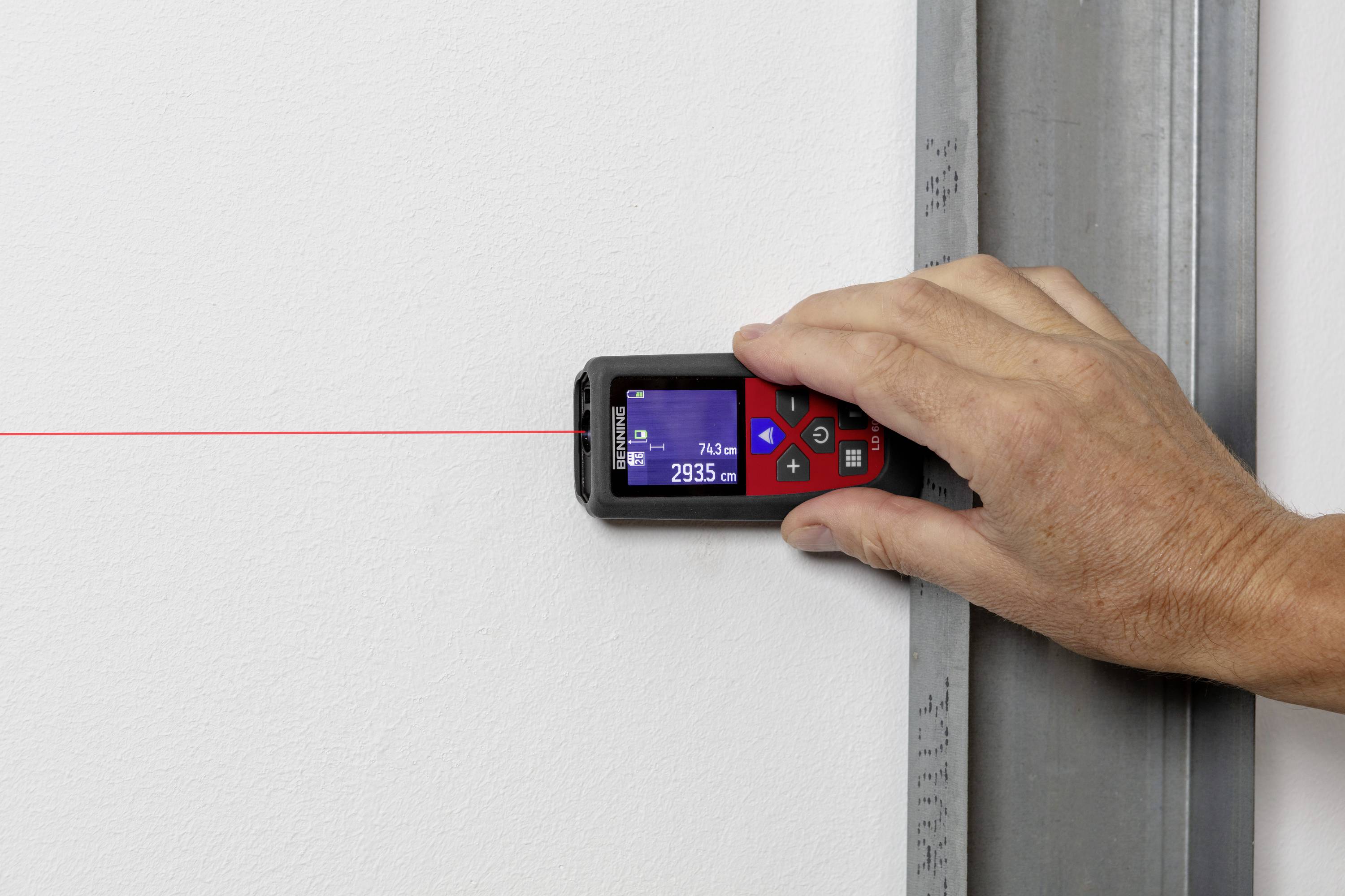 Hand hält einen digitalen Laser-Entfernungsmesser an der weißen Wand. Ein roter Laserstrahl zeigt die gemessene Entfernung von 293,5 cm an.