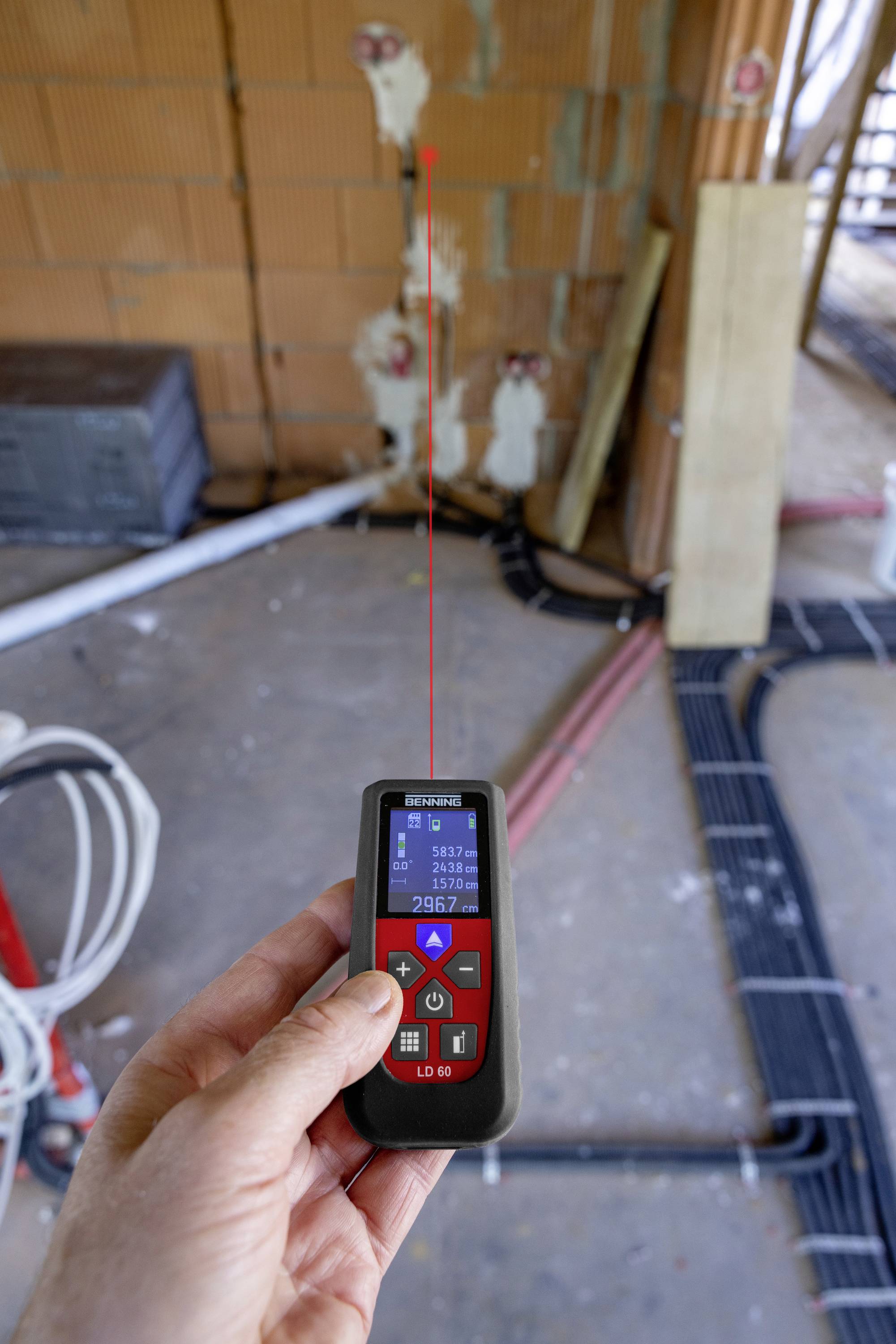 Eine Hand hält ein Laser-Entfernungsmesser in Raum bei Bauarbeiten, mit einem roten Laserstrahl, der auf eine Wand zielt.