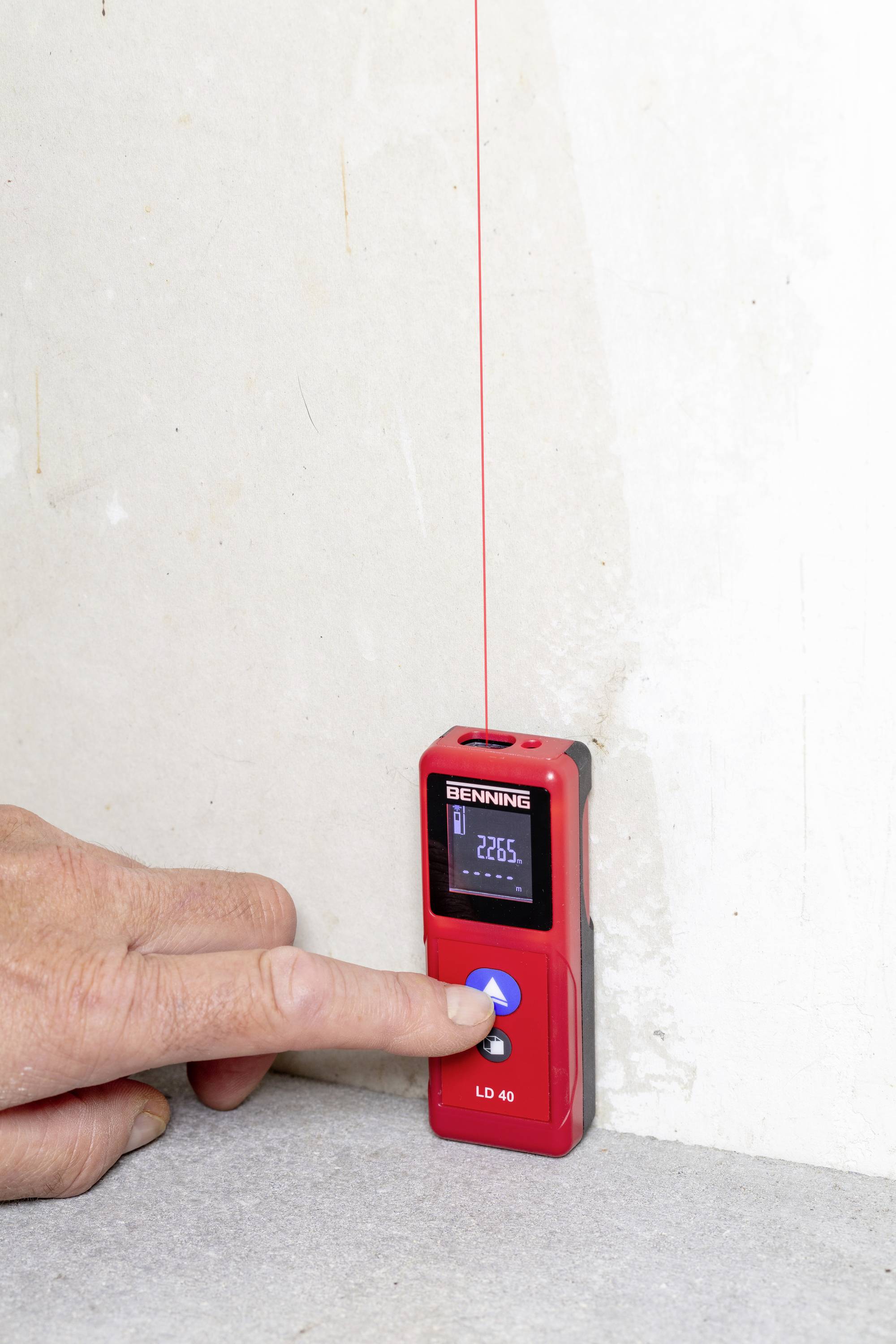 Eine Person hält ein rotes Laser-Distanzmessgerät an eine Wand und drückt einen Knopf. Ein roter Laserstrahl ist sichtbar.