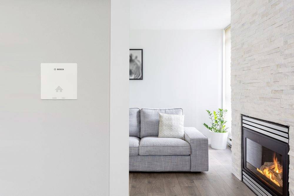 Wohnzimmer mit grauem Sofa, modernem Kamin und grünem Pflanzenakzent. An der Wand ist ein kleines, unauffälliges Gerät installiert.