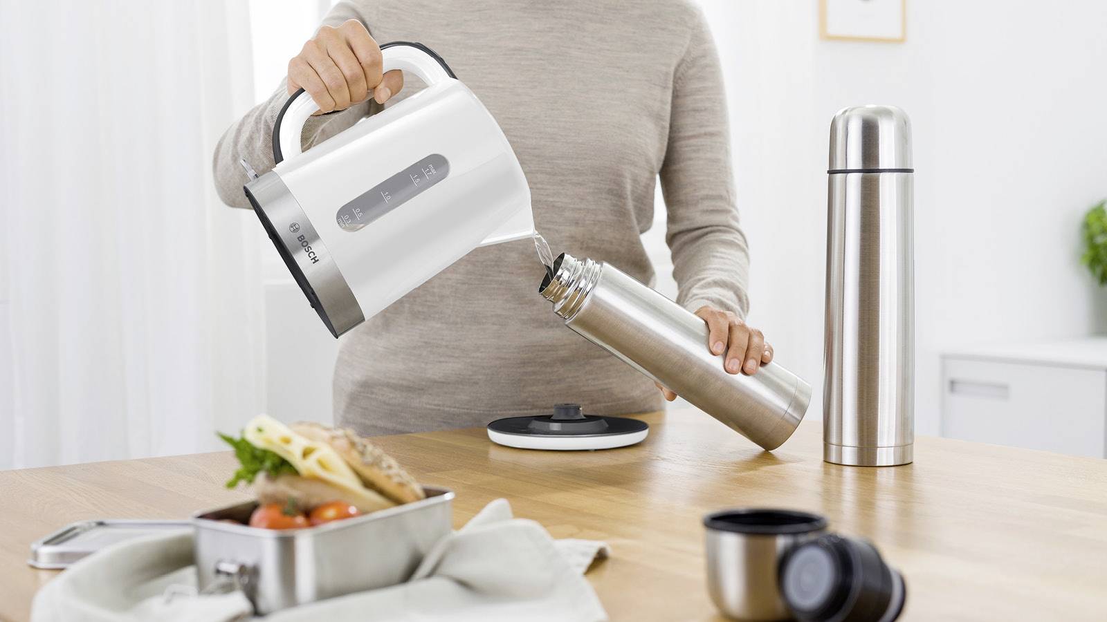 Eine Person füllt heißes Wasser aus einem Wasserkocher in eine Thermoskanne. Im Vordergrund steht eine Lunchbox mit Gemüse und Brot.